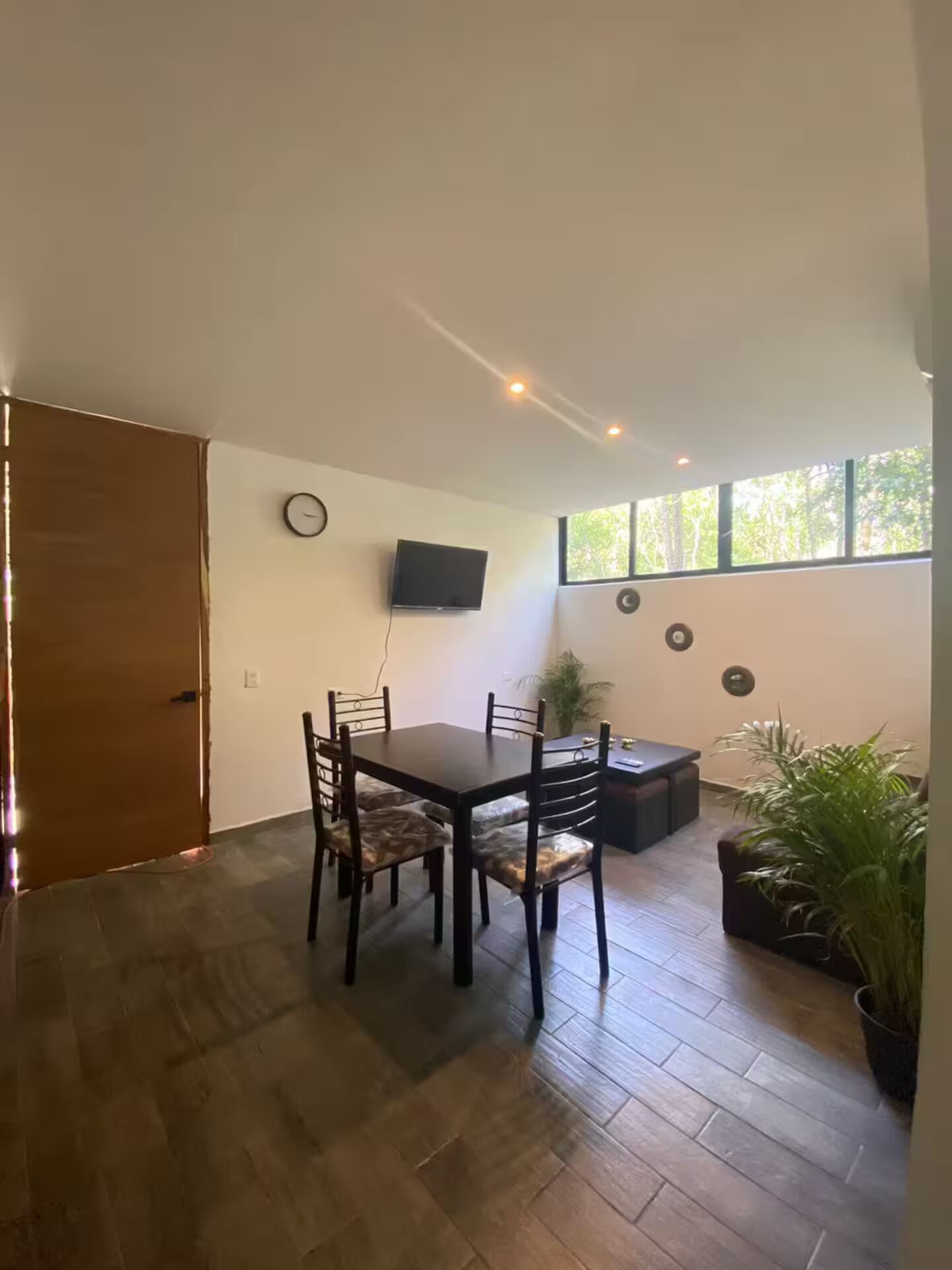 Un coin repas avec une table et des chaises en bois, un téléviseur fixé au mur et une fenêtre donnant sur des arbres.