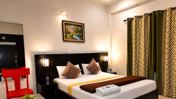 Habitación de hotel con cama, mesitas de noche, una silla roja, una mesa de vidrio con una planta, un cuadro en la pared y cortinas verdes.