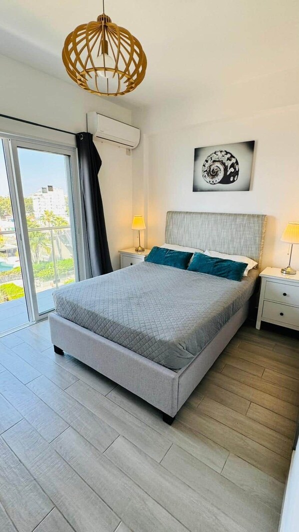 A modern bedroom with a large bed, two bedside tables, a ceiling light fixture, a wall-mounted air conditioner, and a window with a view of buildings.