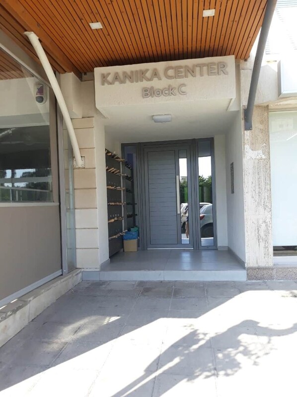 L’entrée d’un bâtiment portant l’inscription « KANIKA CENTER, bloc C », avec un plafond en bois, une porte en verre et un sol carrelé.