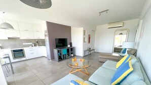 Un saló modern amb un sofà, una taula de centre de vidre i una zona de cuina amb armaris i una televisió.