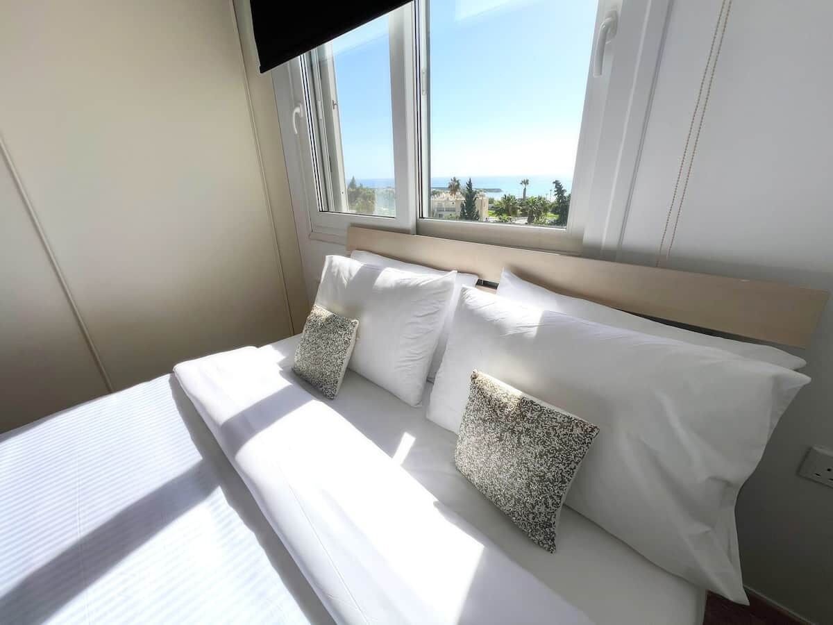 Un letto con lenzuola bianche e due cuscini a fantasia, una finestra vista mare e una testiera in legno.