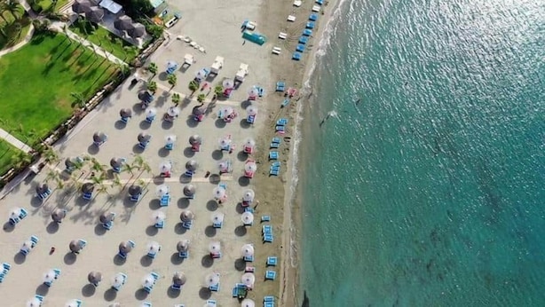 Oversigtsbillede af en strand med rækker af parasoller og strandstole, en sandstrand og et roligt hav.