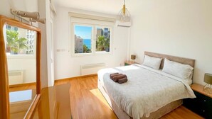A bedroom with a bed, a window, a ceiling light, and a view of buildings and palm trees.