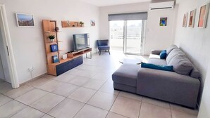 Una sala de estar moderna con un sofá gris, un televisor sobre un mueble de madera, un balcón con puerta corredera y vistas a la ciudad.