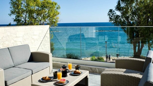 Uma varanda com móveis de vime, uma mesa de vidro com itens de café da manhã e vista para o mar.