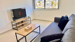 Una sala de estar moderna con televisor, una mesa de centro de madera, un sofá gris y una ventana con vista a la ciudad.