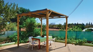 Uma pérgola de madeira com mesa e cadeiras por baixo, rodeada por vegetação e vista para um corpo de água.