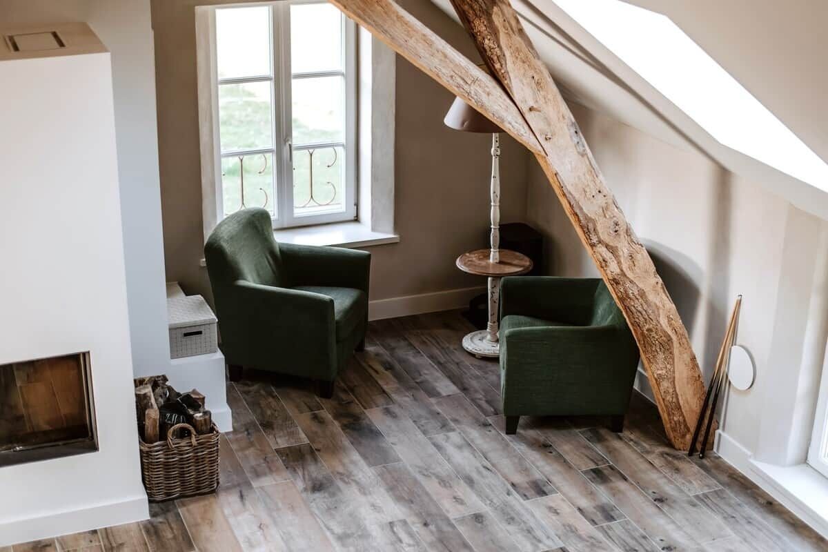 Et loftrum med bjælkebeloft, pejs, to grønne lænestole og et rundt bord.