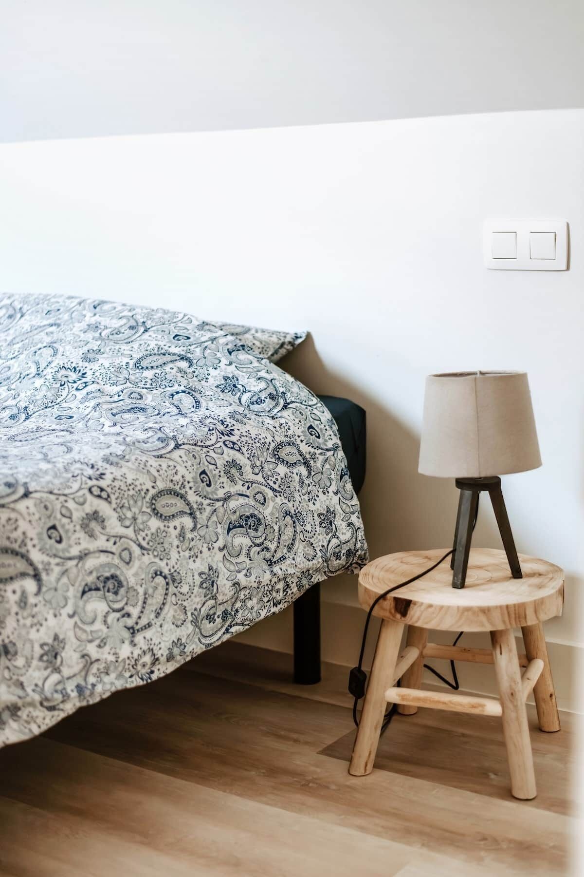 Uma cama com colcha estampada, mesa de cabeceira com abajur e uma tomada na parede.