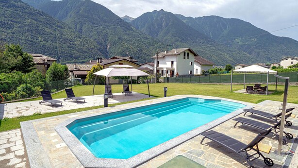Ein Schwimmbad mit Liegestühlen, einer überdachten Terrasse und Häusern im Hintergrund mit Bergen.