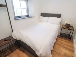 Un dormitorio con una cama, una mesita de noche, una lámpara, una ventana y un control remoto sobre un piso de madera.
