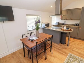 A modern kitchen with a dining area, a TV mounted on the wall, and a window with blinds. - Gews Farm Lodge (Penzance)