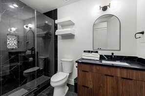 A modern bathroom with a glass shower enclosure, a white toilet, a wooden vanity, and a mirror with lights. - Stylish Retreat Near Downtown Houston w/ Rooftop Views (Houston)