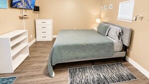 Ein Schlafzimmer mit Bett, Deckenventilator, Fernseher, Kommode und Fenster mit Jalousien.