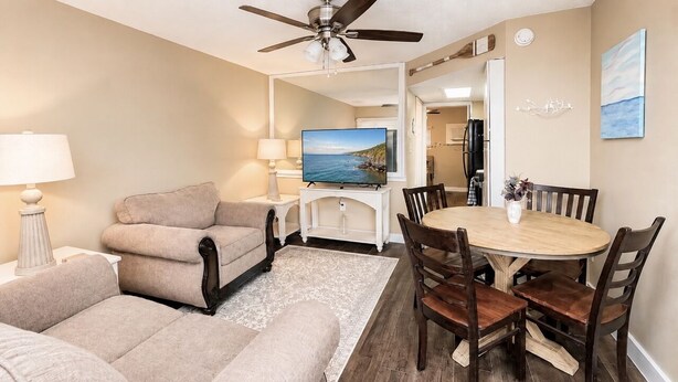 Ein Wohnzimmer mit Sofa, Fernseher, Esstisch mit Stühlen, Deckenventilator und Blick aufs Meer.