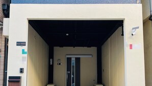 L’entrée d’un immeuble, avec une boîte aux lettres, des poubelles et une porte au-dessus de laquelle est affichée une enseigne.