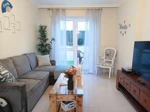 Een woonkamer met een bank, een bijzettafeltje met bloemen, een televisie op een kast en een stoel met een naamplaatje.