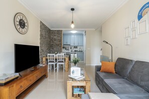 A living room with a flat-screen TV, wooden coffee table, grey sofa, stone wall, and a dining area with white chairs.