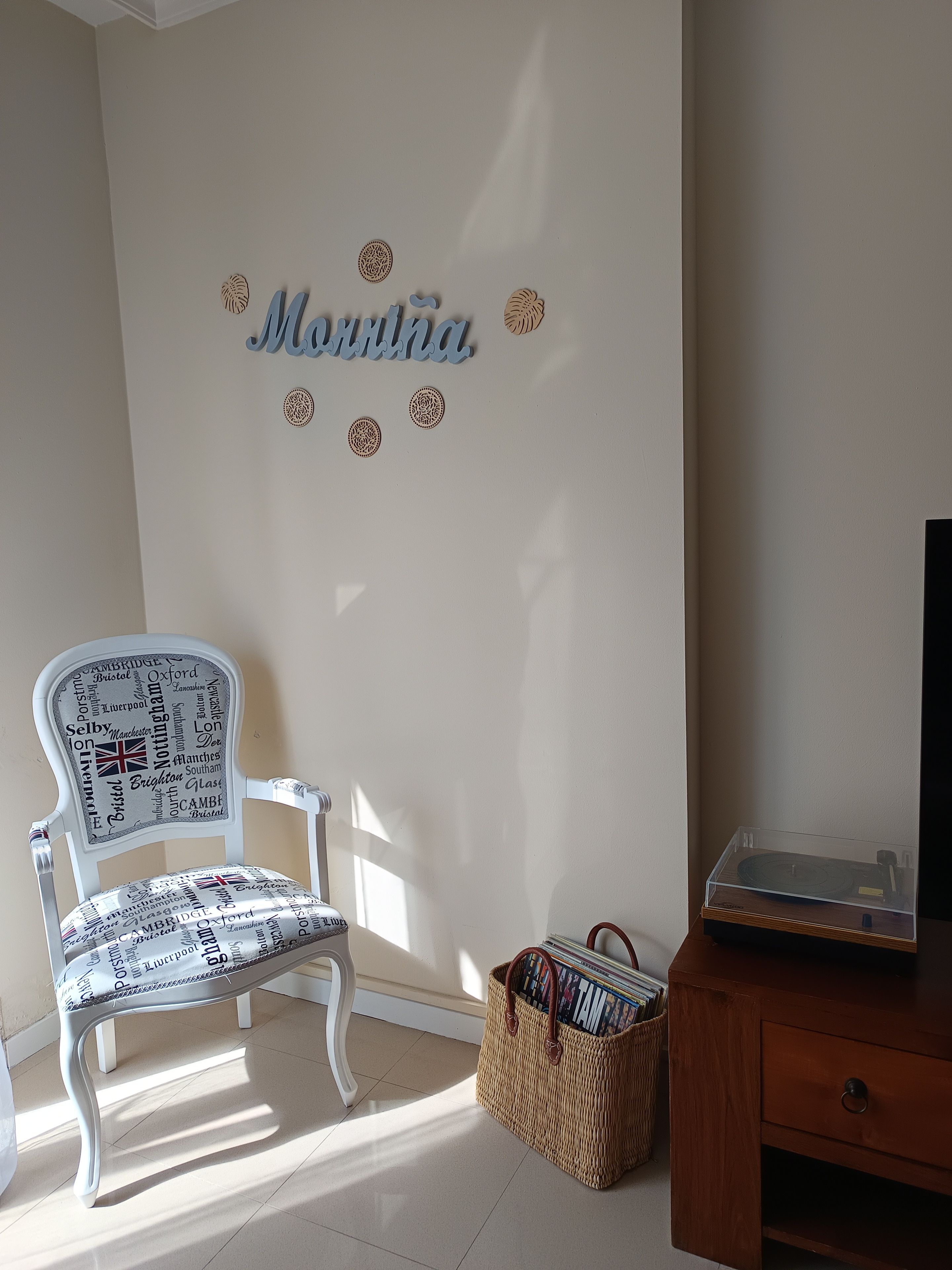 Una habitación con una silla blanca con una placa, una mesita de madera con un televisor y una cesta de mimbre.