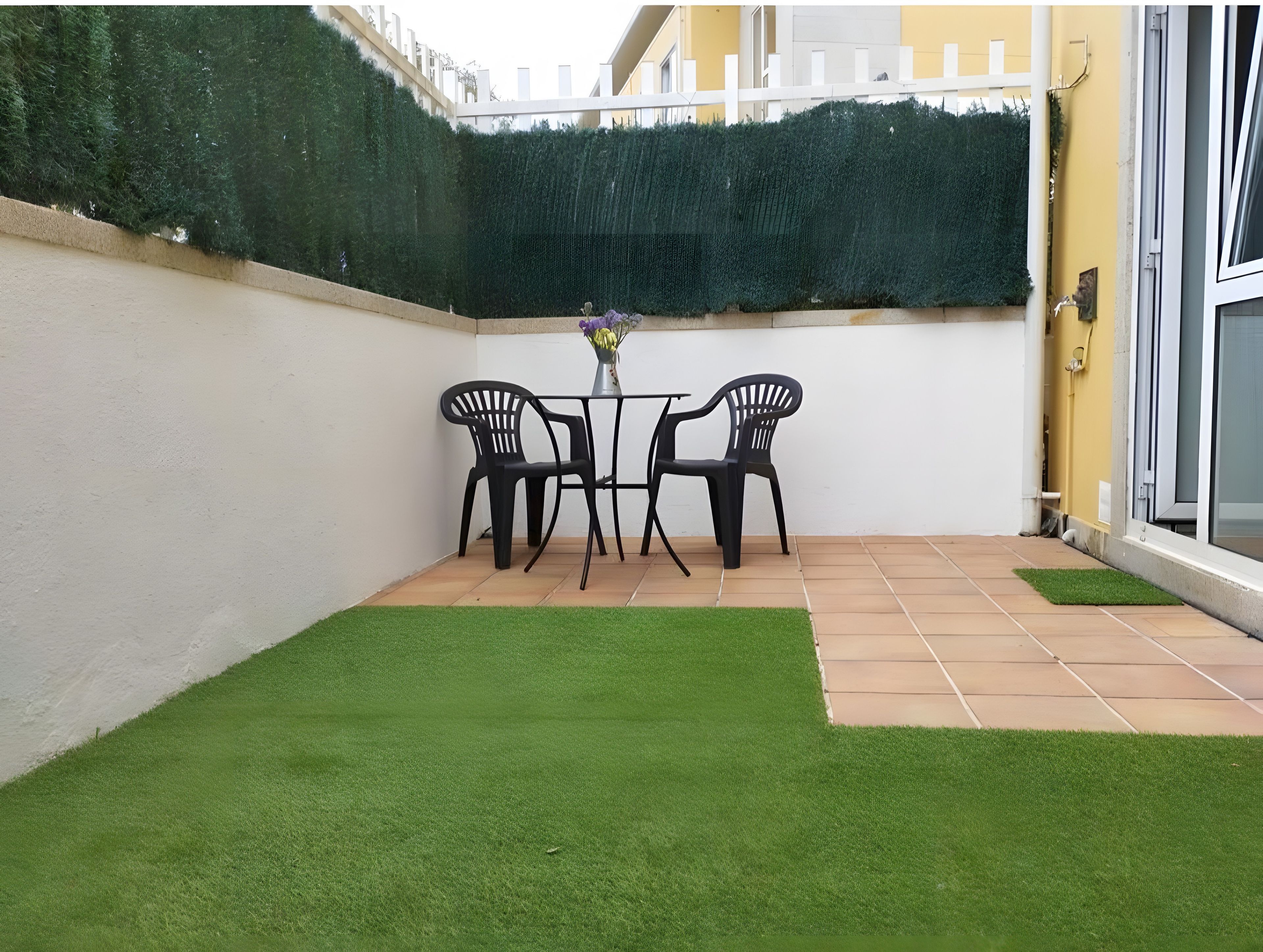 Ein kleiner Patio-Bereich mit Kunstgrras, einem Tisch und zwei Stühlen, begrenzt durch eine Hecke.