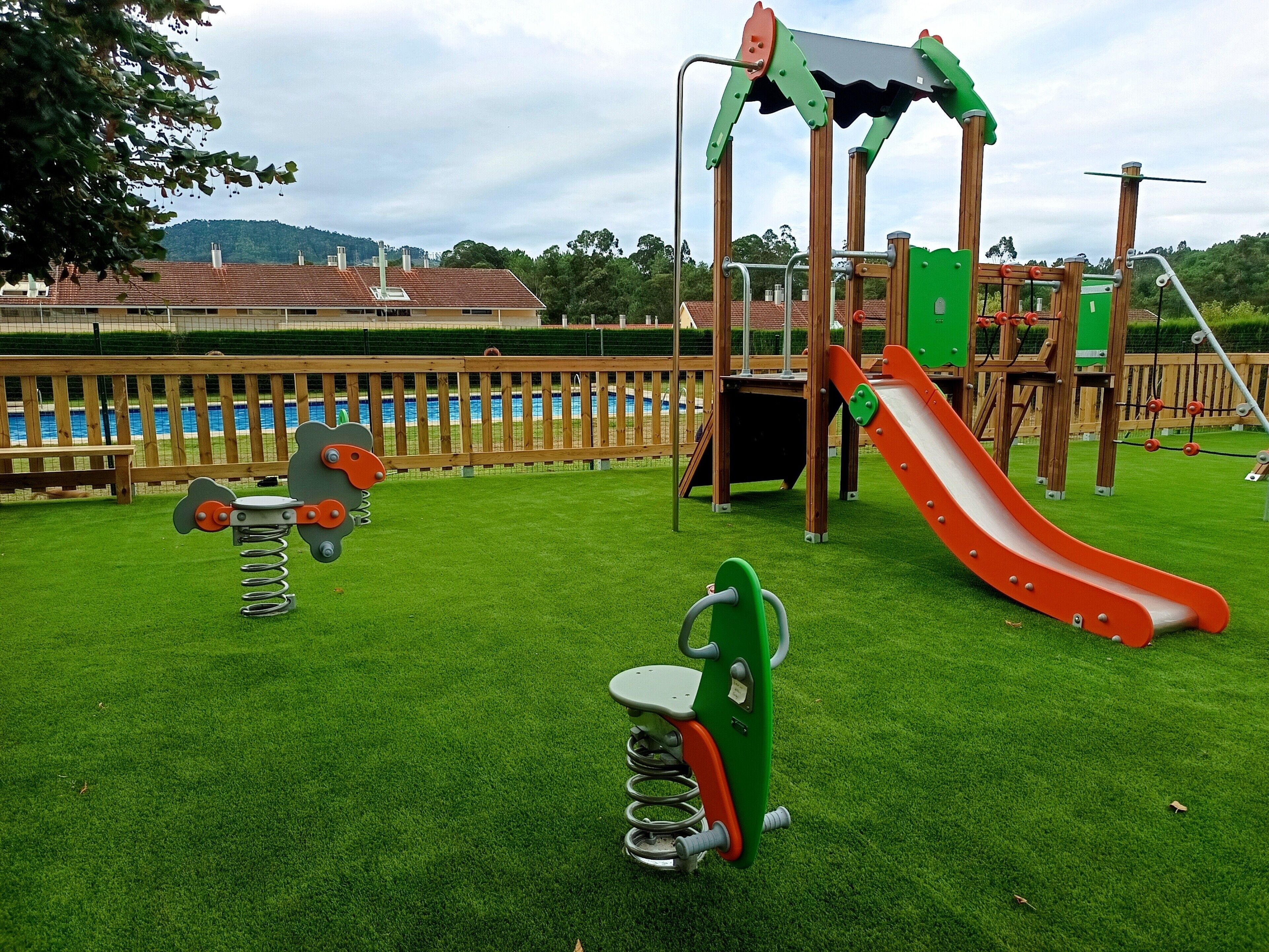 Un parque infantil con un tobogán y columpios, rodeado por una valla de madera y una piscina al fondo.