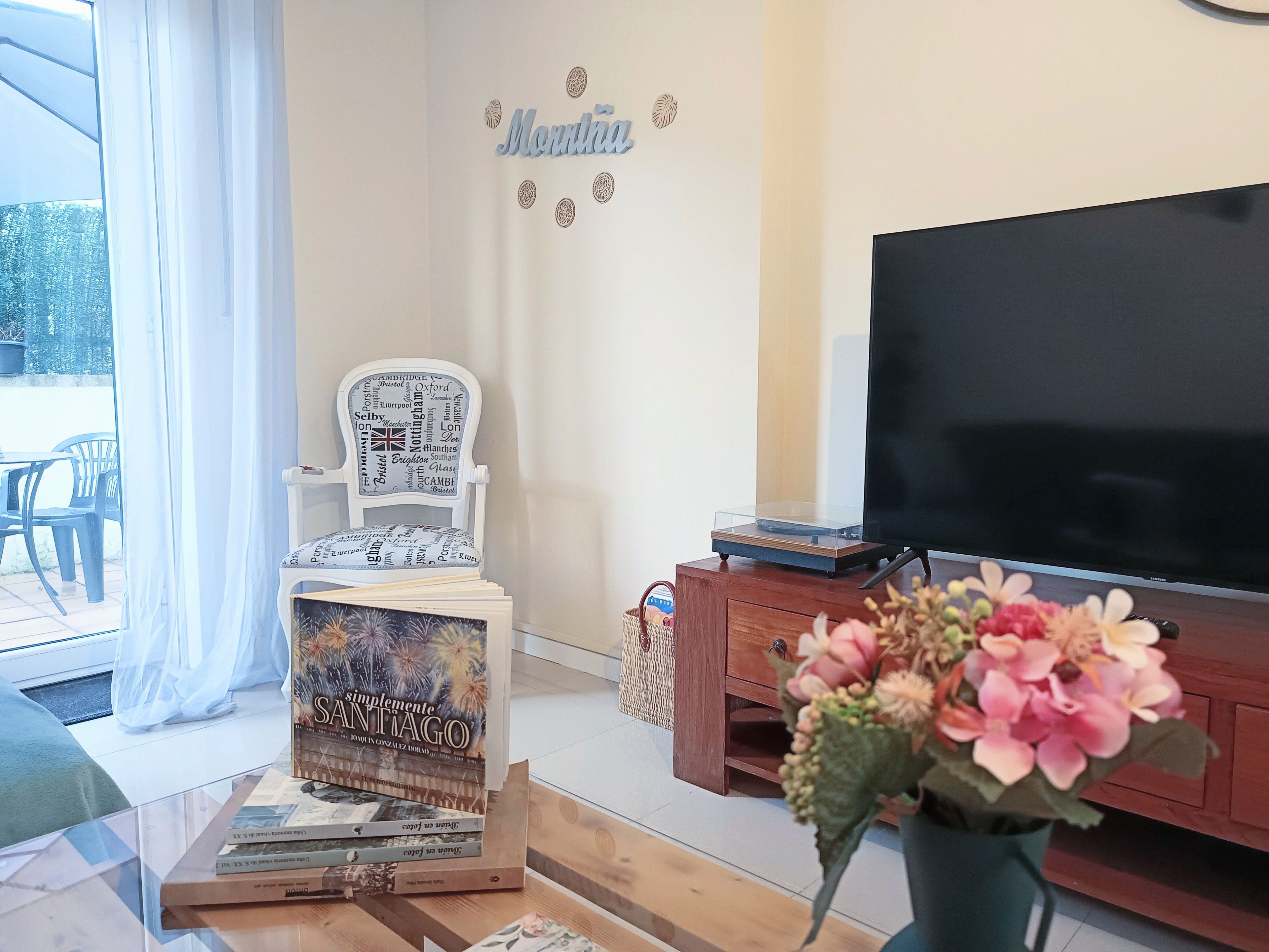 A living room with a TV, a white chair with a map, a bookshelf, and a vase with flowers.