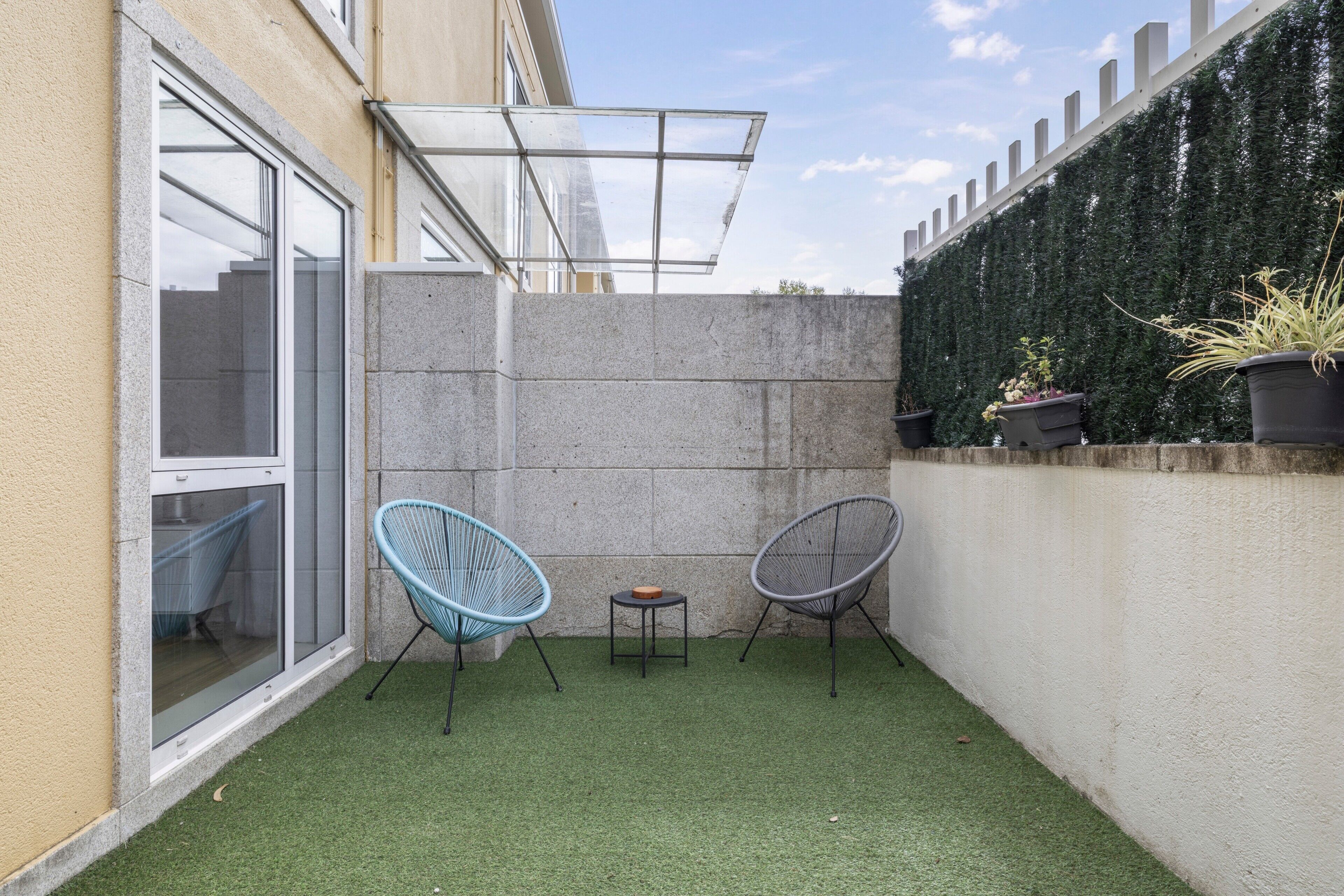 Un pequeño patio al aire libre con césped artificial, dos sillas, una mesita y un ventanal de vidrio que da a un edificio.
