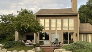 Una casa moderna de dos pisos con un amplio ventanal, un patio con mobiliario exterior y un sendero de piedra que conduce a la entrada.