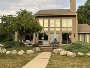 Une maison moderne à deux étages, dotée d’une grande baie vitrée, d’un patio avec des meubles d’extérieur et d’un chemin de pierre menant à l’entrée.