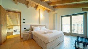 Ein Schlafzimmer mit einem großen Bett, Holzbalkendecke, einem Nachttisch, einer Kommode und Blick ins Freie durch eine Schiebetür.