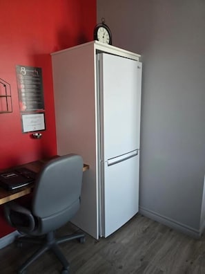 A compact room with a white refrigerator, a desk with a chair, and a clock on top of the fridge.