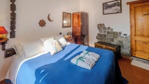 A bedroom with a bed, a nightstand, a wooden wardrobe, a stone fireplace, and a wall-mounted shelf.