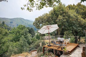 Eine Terrassenanlage im Freien mit Tisch und Stühlen, einem weißen Sonnenschirm und Blick auf einen bewaldeten Hang.