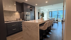 A modern kitchen with dark cabinetry, a marble island, and a dining area with bar stools. - Luxury Lakefront Retreat | Beach, Pool, Hot Tub & Stunning Sunsets (Kelowna)