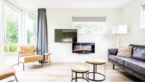 A modern living room with a sofa, two round tables, a television, and a large window with curtains. - Gem With Dune Views for 4 (Vlissingen)
