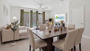 Un comedor moderno con una mesa grande, sillas, un ventilador de techo y una pantalla de televisión mostrando un logotipo.