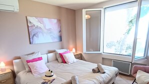 A spacious bedroom with a large bed, a bedside table, a nightstand, a window with a view, and a ceiling light. - Air-Conditioned Mas Juan Villa with Pool and Large Garden in a Quiet Setting, Panoramic Ocean View (Bormes-les-Mimosas)