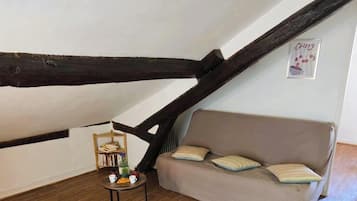 Een knus vertrek met een schuine kap, een houten boekenplank, een ronde tafel met een plant en een bank met gestreepte kussens.