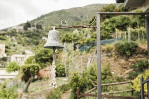 A bell hanging on a metal post with a chain, set against a backdrop of a hilly landscape with buildings and greenery. - Camere Giuliano Basso - Little Suite Sofia  (Vernazza)
