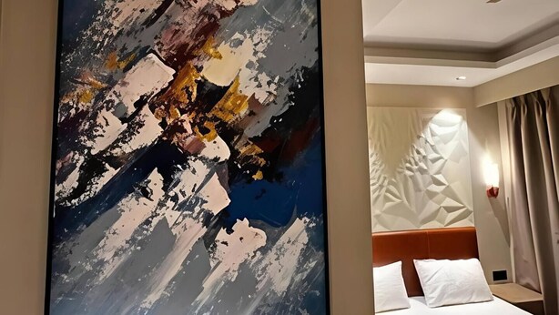 Une chambre d’hôtel avec un grand tableau abstrait au mur, un lit avec du linge de maison blanc et un luminaire moderne.