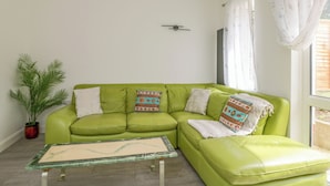 Un sofá verde con cojines estampados, una mesita, una planta en maceta y una ventana con cortinas.