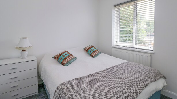 Un dormitorio con cama, dos almohadas, una mesita de noche con lámpara, una ventana con persianas y un radiador.