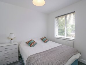 A bedroom with a bed, two pillows, a bedside table with a lamp, a window with blinds, and a radiator. - 167 Leybourne Avenue (Bournemouth)