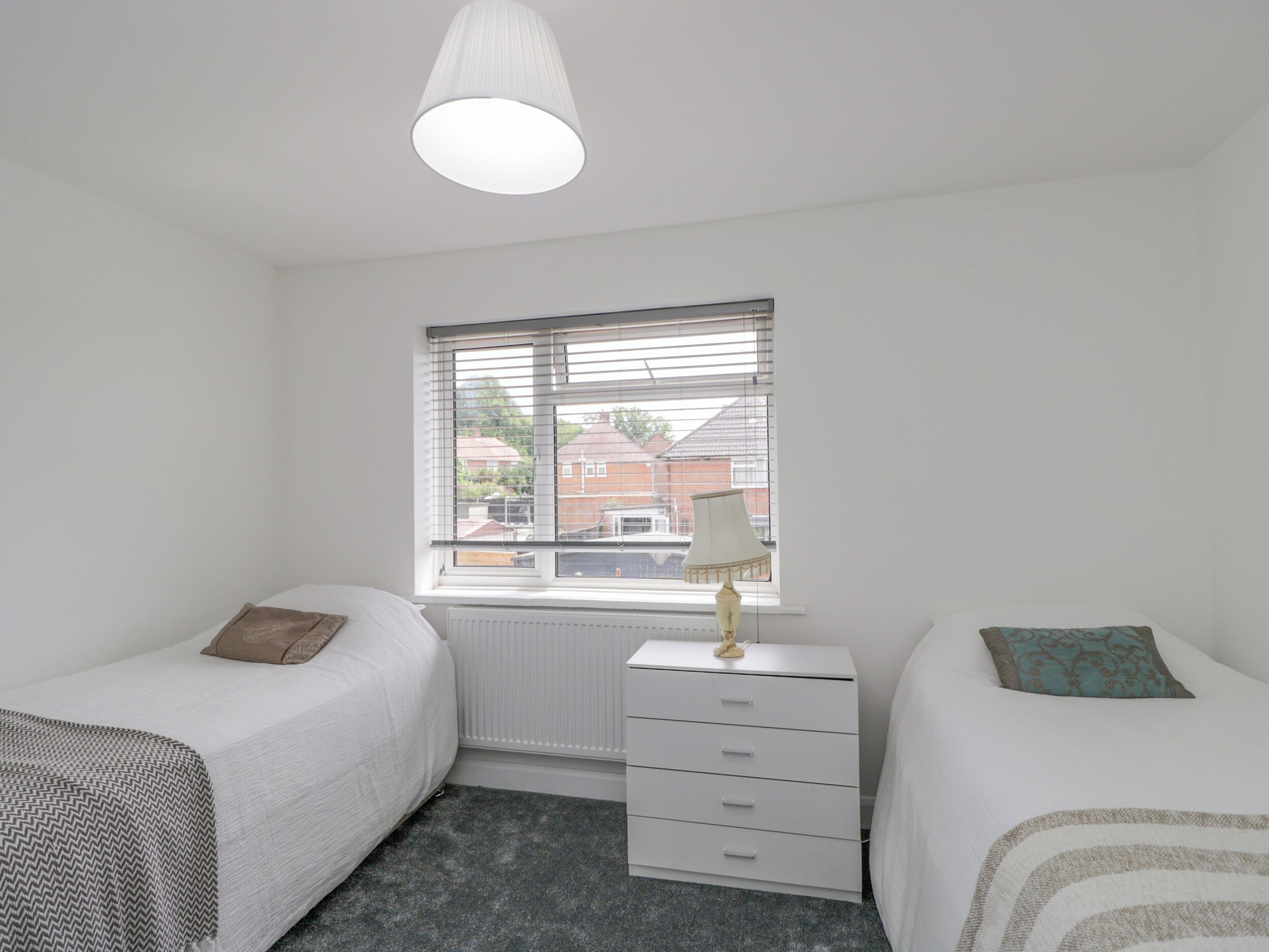 Une chambre avec deux lits simples, une commode blanche, une fenêtre avec des stores et un luminaire au plafond.