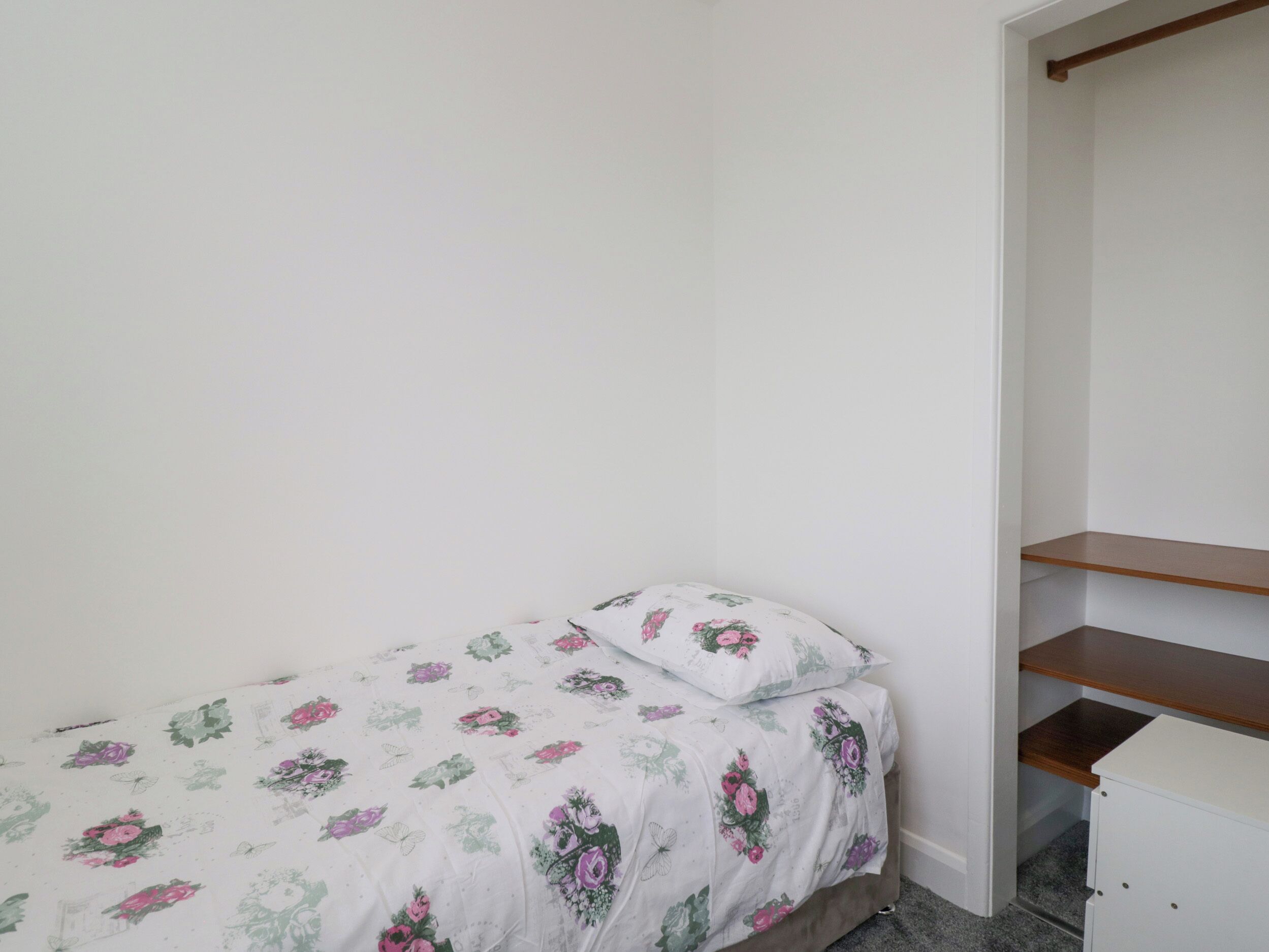 A small, single-bed room with a floral bedspread and a wooden shelf unit.