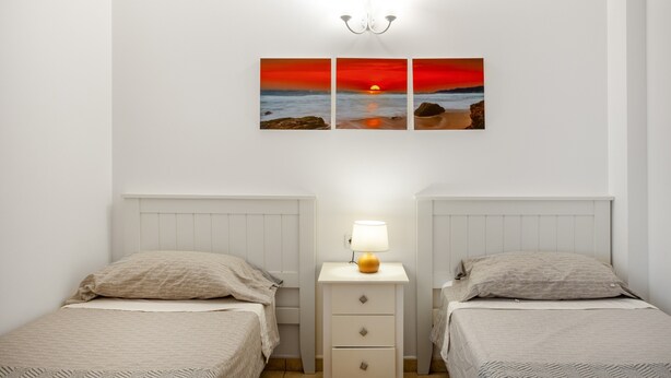 Et soveværelse med to senge, et natbord med lampe, og et billede på væggen af en solnedgang over havet.