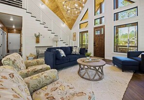 A spacious living room with a dark blue sofa, patterned armchairs, a round coffee table, and a staircase leading to an upper level. - NEW! Heated Pool, Hot Tub, Pickleball, Games, EV (Broken Bow)