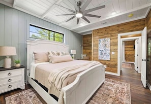 A bedroom with a bed, nightstands, a ceiling fan, a window, and a wooden wall. - NEW! Heated Pool, Hot Tub, Pickleball, Games, EV (Broken Bow)
