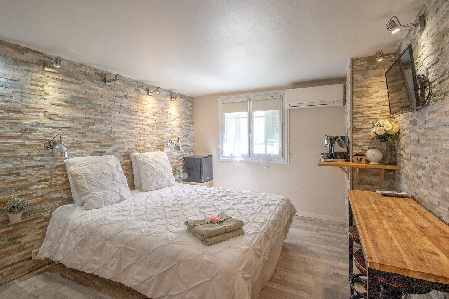 Un dormitorio con pared de piedra, una cama con colcha, un escritorio de madera y un televisor montado en la pared.